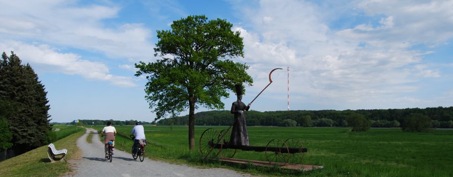 Urlaubsregion Höhbeck Elbe - Titelbild: Blick vom Elbdeich auf den Höhbeck mit Funkturm. Im Vordergrund eine Bronzeskulptur eines mythischen Fährmanns.