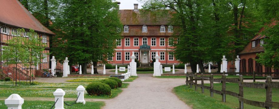 Urlaubsregion Höhbeck Elbe - Titelbild: Barockschloss Gartow (rot gestrichen mit weißen Fenstereinfassungen und herrschaftlicher Auffahrt) mit hinter Bäumen versteckten Seitenflügeln. Im Vordergrund weiße Tordurchfahrt und geschotterte Auffahrt.