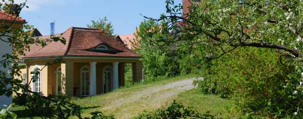 Eingeschossiges okkafarbenes Nebenggebäude der Burg Lenzen mit weißen bodentiefen Fenstern und flachem Ziegeldach. Das Dach wird im Vorraum von zwei weißen Säulen gestützt.