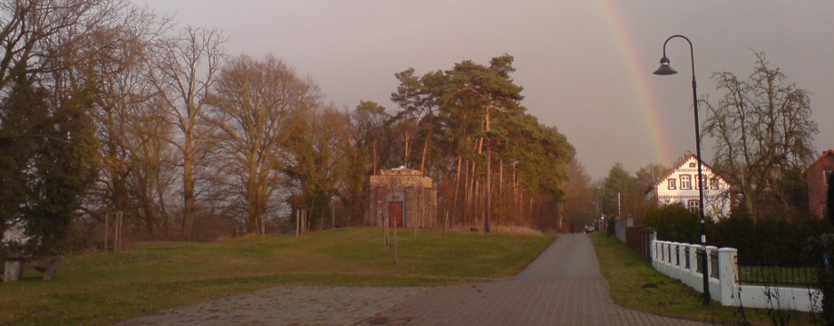 Urlaubsregion Höhbeck Elbe - Titelbild: Gedenkhalle aus Feldsteinen in Form eines Mausoleums  und weißes Schifferhaus am Hochufer der Elbe in Vietze. Im Hintergrund ein Regenbogen.